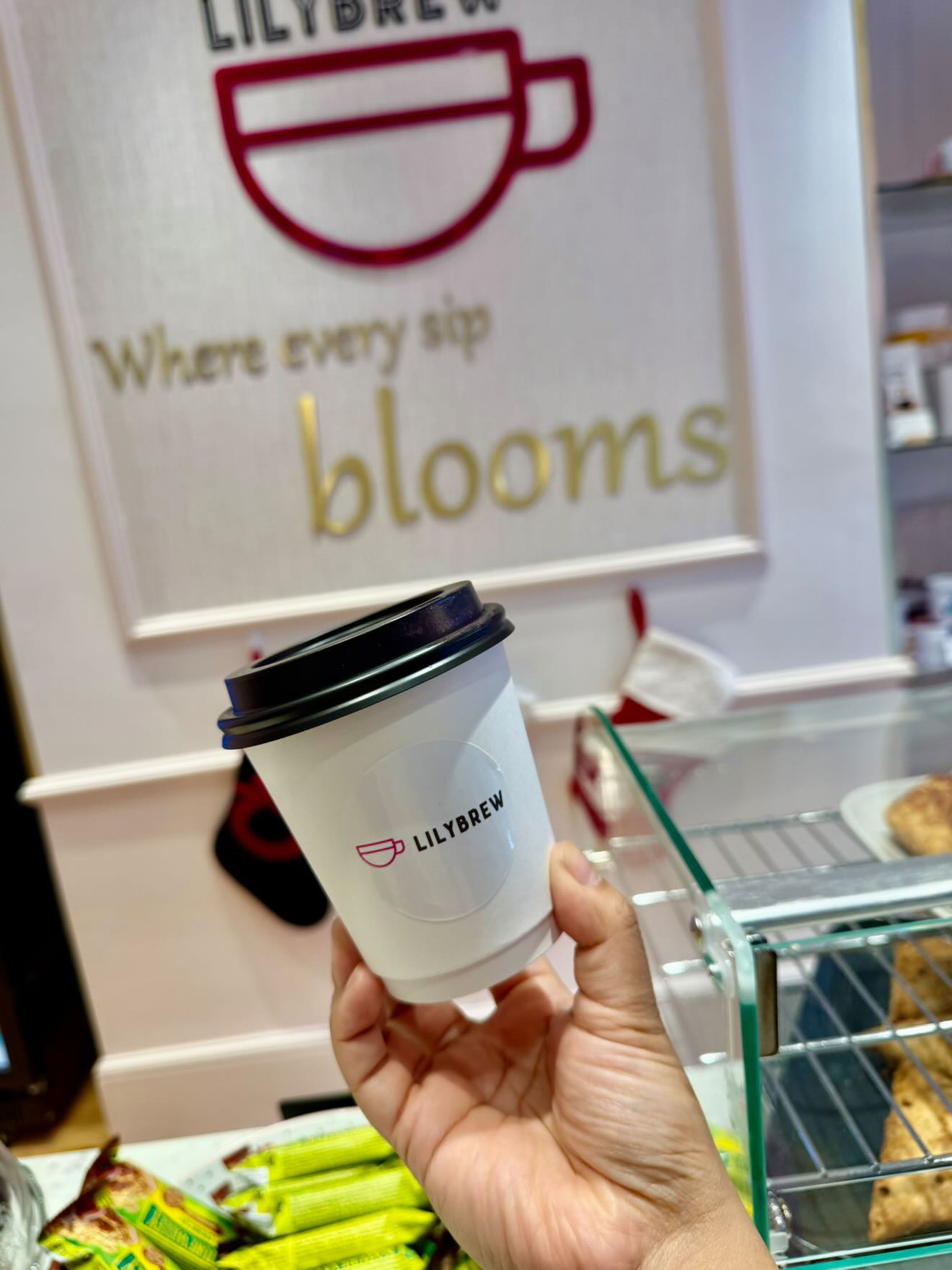 A takeaway coffee cup from Lily Brew Jesmond, white cup featuring the logo and black plastic top